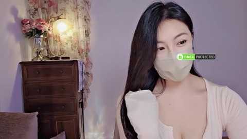 Media: Video of an East Asian woman with long black hair, wearing a white face mask, a beige top, and a white glove, standing in a softly lit room with a floral-patterned wall and wooden dresser.