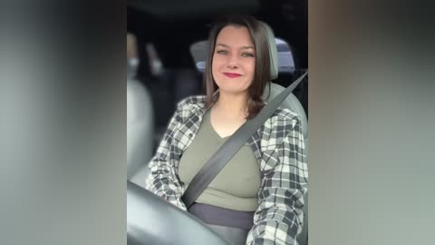 Media: Video of a smiling young woman with light skin and shoulder-length brown hair, wearing a gray shirt and plaid shirt, driving in a car.