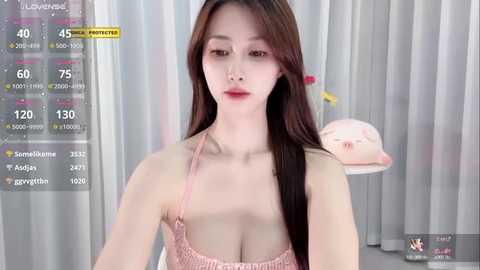 Media: A video of an East Asian woman with long, straight brown hair, fair skin, and a slender physique. She wears a light pink bralette, revealing her ample cleavage. The background features a modern, minimalist room with white curtains.