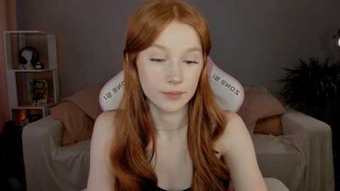 Media: Video of a young, fair-skinned redhead woman with long, wavy hair, wearing a black tank top, sitting in a gaming chair with a \"The Backroom\" logo, in a cozy, dimly lit living room.