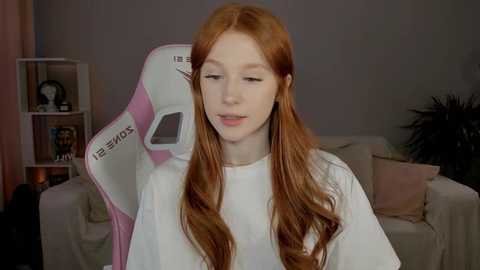 Media: Video of a young, fair-skinned, red-haired woman with wavy hair, wearing a white top, sitting in a pink and white gaming chair. Background features a beige sofa, a potted plant, and a bookshelf.