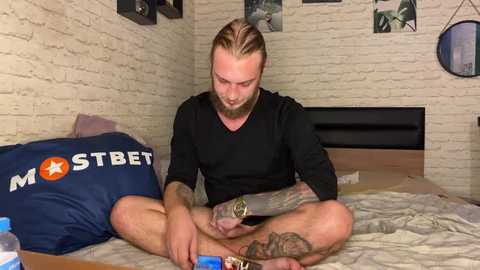 Media: Video of a tattooed, bearded man with long hair, wearing a black shirt, sitting cross-legged on a bed with a blue pillow. He's focused on a smartphone, surrounded by a minimalist, beige-brick-walled room.