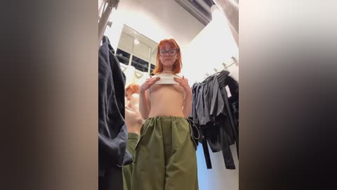 Media: Video of a topless woman with short red hair, glasses, and green pants, lifting her shirt in a dimly lit dressing room.