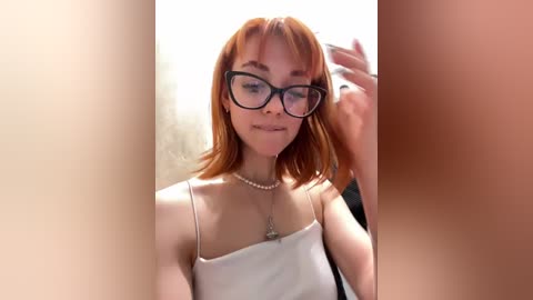 Media: A video of a fair-skinned woman with short, red hair, wearing large black-rimmed glasses and a white spaghetti-strap top, adjusting her hair in a mirror, blurred background.