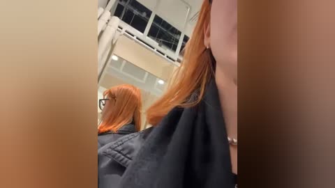 Media: A video of a woman with long, straight red hair, wearing black-rimmed glasses and a dark robe, captured from behind in a modern, well-lit room with reflective surfaces.