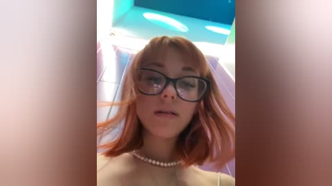 Media: Video of a young woman with shoulder-length, light brown hair, wearing black-rimmed glasses and a pearl necklace. She stands indoors, surrounded by a blue and white tiled bathroom with a circular light fixture above.