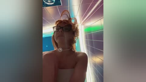 Media: A video of a woman with curly red hair, wearing dark-rimmed glasses and a pearl necklace, standing in a futuristic, neon-lit room with tiled walls and a circular light fixture.