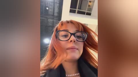 Media: A close-up video of a young woman with straight, shoulder-length, reddish-brown hair and large, black-rimmed glasses. She wears a black coat and a pearl necklace. The background features a modern, indoor setting with white walls and a large window.