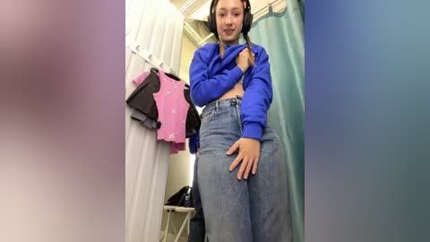 Media: Video of a young Asian woman with medium build and fair skin, wearing a blue sweater and high-waisted jeans, standing in a dimly lit dressing room with pink and green shower curtains.