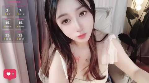 Media: A video of an East Asian woman with long, straight black hair, fair skin, and a slender build, wearing a white sleeveless top. She's sitting on a bed, with a green plant and a lamp in the background.