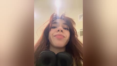 Media: Video of a young woman with long, wavy auburn hair, wearing black over-ear headphones, making a pouty face in a dimly lit room with cream-colored walls.