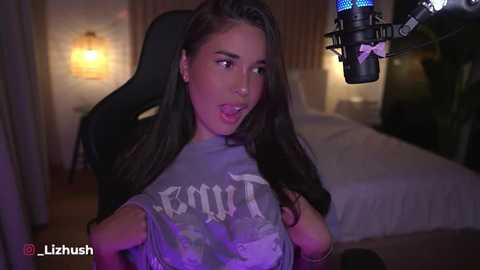 Media: Video of a young woman with long dark hair, wearing a gray T-shirt, in a dimly-lit bedroom, holding a microphone, with a black gaming chair in the background.