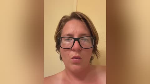 Media: A video of a fair-skinned, middle-aged woman with shoulder-length, light brown hair and black-framed glasses, looking serious. She is topless, set against a plain, light beige background.