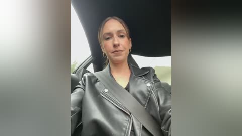 Media: Video of a Caucasian woman with light skin and brown hair in a ponytail, wearing a black leather jacket with a shoulder strap, seated in a car.