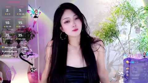 Media: Video of an East Asian woman with long black hair, wearing a black top, sitting in a room with a green plant, pink fairy decorations, and a digital calendar.