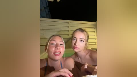 Media: Video of two topless young women with light skin and blonde hair, sitting in a hot tub at night, illuminated by warm lights.