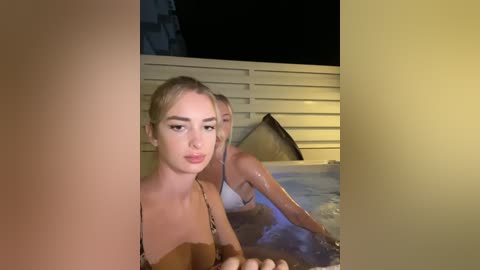 Media: A video shows two blonde women in bikinis, one kissing the other, in a dimly lit, outdoor hot tub; the background features a wooden fence and a dark sky.