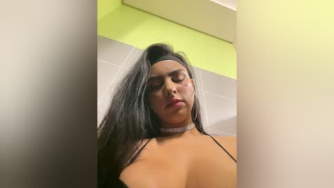 Media: Video of a young, Latina woman with long black hair, wearing a black spaghetti strap top and a white choker necklace, standing in a brightly lit bathroom with green and white tiled walls.