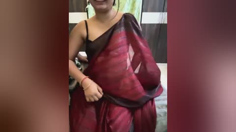 Media: Video of a South Asian woman wearing a traditional red sari with black borders, adjusting the fabric with her right hand. Background features a blurred room with green and brown elements.