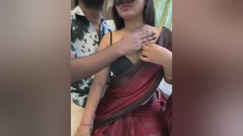 Media: A video showing a man with a beard, wearing a patterned shirt, lifting a woman's black bra strap, revealing her red sari and medium-sized breasts.