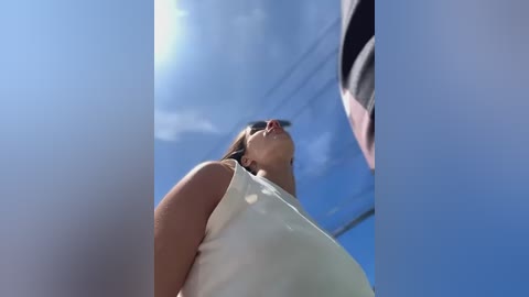 Media: Video of a woman in a white tank top, looking upwards with her mouth open, against a clear blue sky with a few clouds.