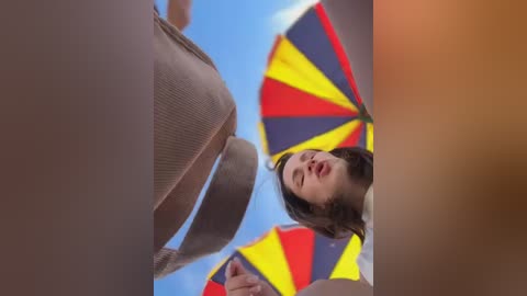 Media: A video of a young woman with short brown hair, wearing a white t-shirt, holding a colorful umbrella with yellow, red, and blue sections, smiling under a bright blue sky.