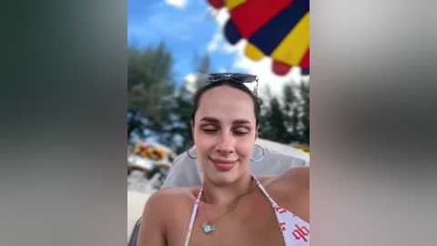 Media: A video of a smiling, light-skinned woman with dark hair, wearing a white bikini with red text and hoop earrings, sitting outdoors under a colorful umbrella.