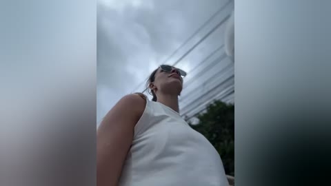 Media: Video of a woman with light skin, dark hair, and sunglasses, wearing a white sleeveless top, standing in front of a modern, glass-walled building with horizontal lines under cloudy skies.