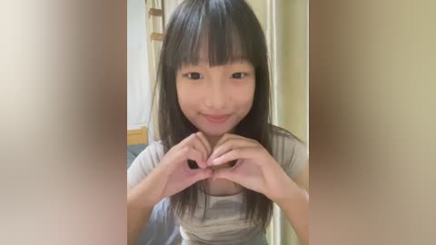 Media: Video of a young Asian woman with long black hair, straight bangs, and light skin, making a heart shape with her hands in front of her chest. She wears a light-colored, short-sleeved shirt. Background shows a beige wall and part of a wooden door.