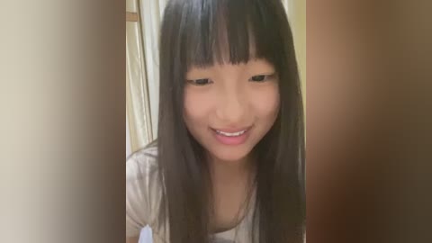 Media: Video of a young Asian girl with long black hair and bangs, smiling, wearing a light gray shirt, standing in a doorway with beige walls.