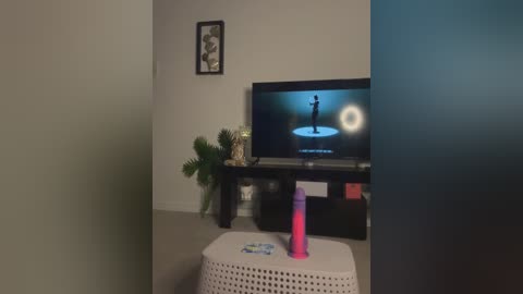 Media: A video shows a living room with a flat-screen TV displaying a blue screen, a white wicker ottoman, and a bright pink dildo on it. The background includes a framed picture and green plants.