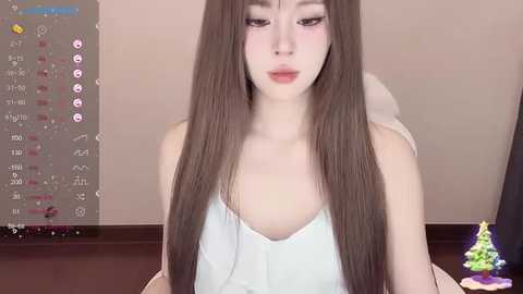 Media: Video of a young Asian woman with long, straight brown hair, wearing a white tank top, sitting indoors. Background features a digital clock and a Christmas tree.