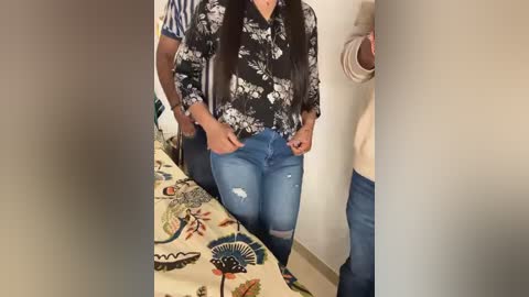 Media: Video of a woman with long dark hair, wearing a floral blouse and distressed jeans, sitting on a colorful bed, being held by a man in a striped shirt.