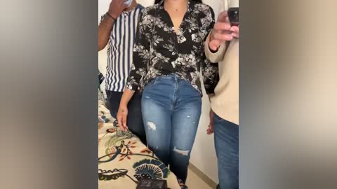 Media: Video of a woman in a black floral blouse and ripped blue jeans, taking a selfie in a room with beige walls and a patterned blanket.