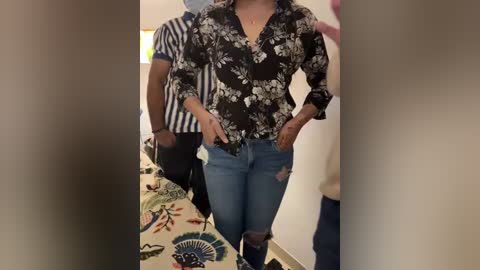 Media: Video of a woman in a black floral blouse and distressed blue jeans, standing in a room with a striped shirt and patterned tablecloth in the background.