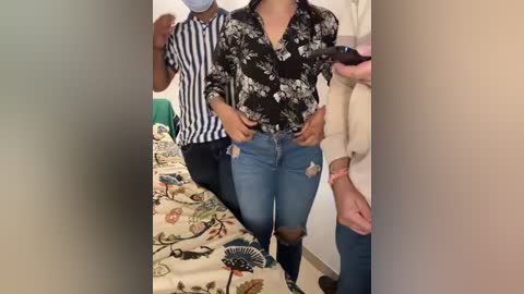 Media: Video of a woman in a floral blouse and ripped jeans, walking in a room with a striped-shirt man, a bed, and a person in a beige sweater.