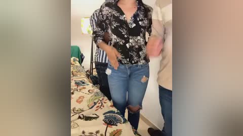 Media: Video of a woman in a floral blouse, blue jeans, and a white jacket, standing in a bedroom with a bed, striped chair, and beige walls.