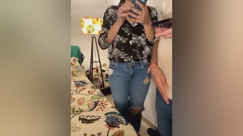 Media: Video of a woman in a floral blouse and distressed jeans taking a mirror selfie in a bedroom with a colorful lamp, bedspread, and white walls.