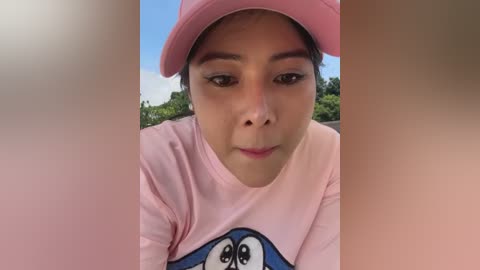 Media: A close-up video of a young woman with medium brown skin and dark brown eyes, wearing a pink hat and shirt featuring a blue cartoon character. Background shows greenery and a bright blue sky.