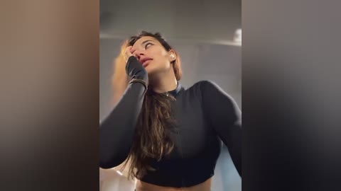 Media: Video of a young woman with long brown hair, wearing a black long-sleeved crop top, standing against a blurred background. She looks contemplative, with one hand touching her hair, and her eyes gazing upward.