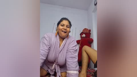 Media: Video of a plump, smiling Latina woman in a lavender robe, sitting on a bed with a red doll and patterned blankets.