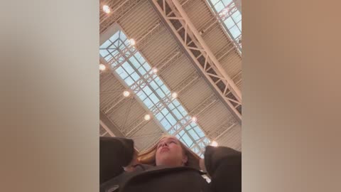 Media: A video captures a woman lying on her back, looking up at a high, glass-paneled ceiling with metal beams and warm lighting. Her blonde hair cascades down, partially covering her face. The scene is bright and open, suggesting an indoor space like a gym or atrium.