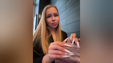 Media: Video of a young blonde woman with fair skin, wearing a black robe, seated at a table in a modern room with grey tiled walls, holding a piece of paper.