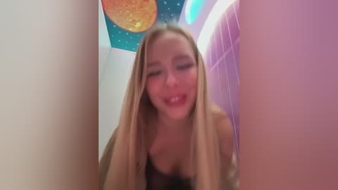 Media: A video of a blonde woman with long hair, wearing a black lace bra, looking playful with her tongue out, in a pink-tiled room with a space-themed ceiling.