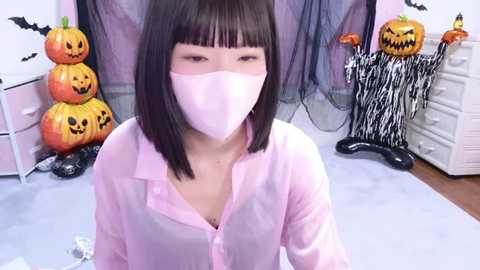 Media: Video of an East Asian woman with straight black hair, wearing a pink mask and shirt, standing in a Halloween-themed room with carved pumpkins and a black spider costume.