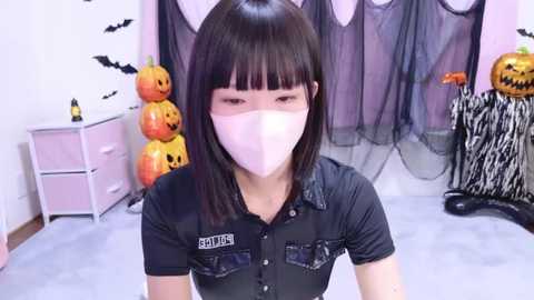 Media: Video of a young Asian woman with straight black hair, wearing a black button-up shirt, pink mask, and a \"SETOUCHI\" name tag. Halloween-themed background with jack-o'-lanterns, bats, and a zebra-print umbrella.