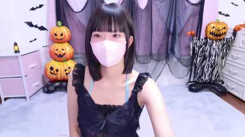 Media: Video of a young East Asian woman with straight black hair and a pink mask, wearing a black lace dress, in a Halloween-themed room with jack-o'-lanterns, bats, and spider decorations.