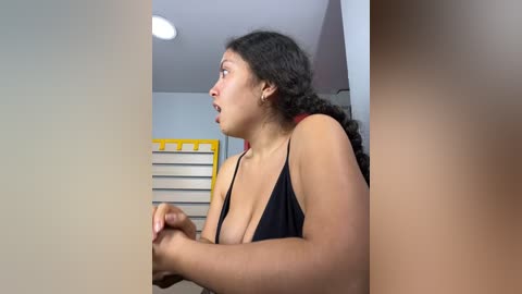 Media: Video of a Latina woman with curly black hair in a black tank top, startled expression, holding a phone, indoors with a yellow-framed window and blinds.