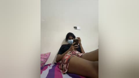 Media: A video shows a young woman with medium brown skin and long dark hair lying on a bed with pink and purple striped sheets, wearing a black t-shirt, floral shorts, and a face mask.