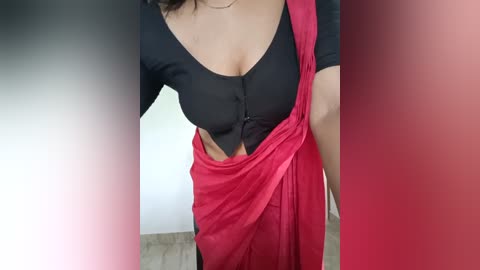 Media: A close-up video of a woman with light skin, wearing a black top with a deep V-neck and a red sari with a matching red blouse, partially draped over her shoulders.